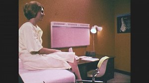 1970s: Woman sits on exam table, wears exam robe. Doctor sits down on chair, talks to patient.