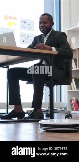 An African American man in a suit sits at a desk, focused on his laptop, while a robotic vacuum cleaner operates on the floor nearby Stock Video Footage - Alamy