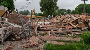 Small loader tractor equipment moves in reverse through a demolition site. Piles of rubble in the foreground of view following destruction of a building. Overcast cloudy skies above.