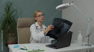 Attractive female doctor in eyeglasses working indoors, looking at x-ray images, taking notes in notepad while sitting at work table in physician office. Common workday of healthcare specialist