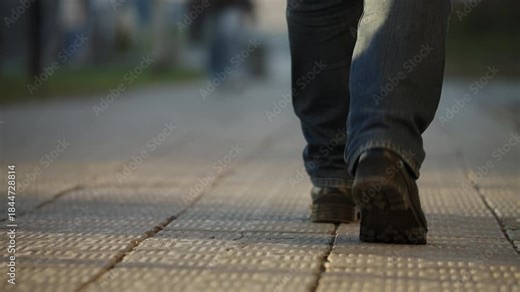 Close-up rear view of a person in jeans and dark shoes walking along a tiled sidewalk in an urban setting, highlighting the steady pace of everyday commute with textured pavement and shallow focus.
