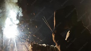 Industrial welder with protection gear welds metal parts in workshop, creating durable joints. Manual arc welding, helmeted worker precision. Construction, metalwork, safety training, Project insight