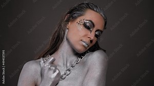 A gorgeous female model with silver body art on her body, bright makeup and silver hair poses for the camera, a chain hanging around her neck. Fashion. Stock Video