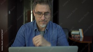 Happy mid adult man working with laptop computer in home office. Businessman smiling, thinking, wearing glasses.