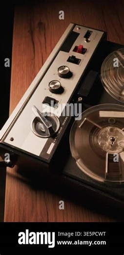 A vintage reel-to-reel tape recorder plays audio on a dark wooden table. Warm, moody lighting highlights the spinning analog reels and metallic dials Stock Video Footage - Alamy