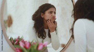 Portrait of sad young dark haired indian woman touching facial skin dissatisfied quality of care products while looking at mirror reflection at home Bad negative skin care treatment beauty concept