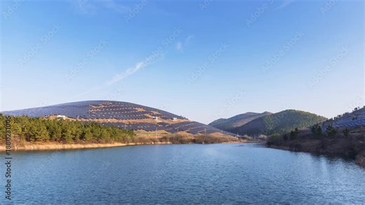 Time-lapse of a mountain solar farm at early morning, photovoltaic panel arrays on hillsides beside a calm reservoir under clear blue sky.