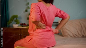 Back view shot of young girl suffering from back pain while sitting on bed during night sleep - concept of cramps, mattresses Problem and strain