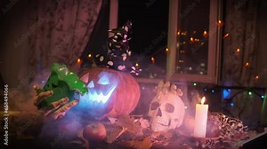 Tattooed male hand throws candy into bucket which is held by Jack-o'-lantern, traditional carved pumpkin rotates and strokes skull against backdrop of autumn during Halloween decorations.