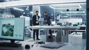 Young Engineer Works with Colleague Robotics Scientist with Laptop Computer. They Collaborate on a Robot Arm Development in an Industrial Startup. Analyzing and Evaluating Technology Systems.