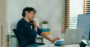 Positive young freelance Asia male use computer and talk on cell phone remote work on desk in living room at home office. Work from house, financial plan, remotely digital nomad job and Gen Z talent.