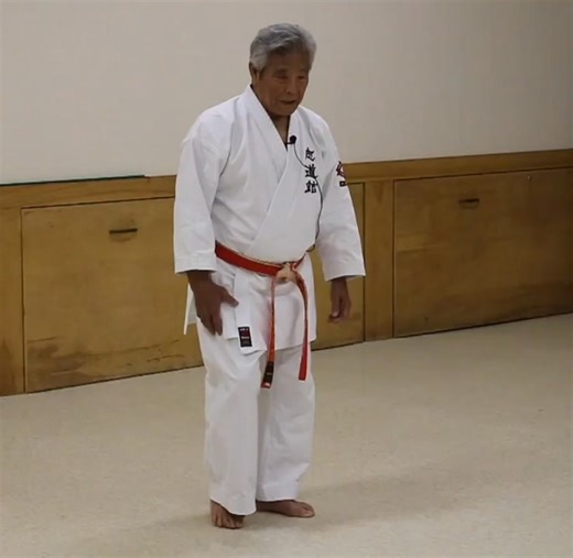 Here Iha Sensei explains the stepping that takes place in Chibana’s kihon kata 1 and 2. The emphasis is on natural motion, and that there is a counter balance between the upper and lower body. The stepping moves through the center of balance in an arcing, half moon manner. The knees do not bend The foot touches not with a heel first, but with the point behind the middle toe, as an outward pushing power engages. As the foot makes contact the legs have tension in the thigh area and on the floor, w
