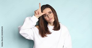 You lose! Brunette young woman making loser gesture, L sign on forehead, teasing and accusing for defeat, expressing disrespect. Indoor studio shot isolated on blue background.