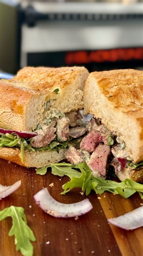 Chef Nathan Lippy brings big heat and bold flavor to the Blackstone Open Flame Grill, searing a ribeye until it forms a rich, crispy crust. He mixes a simple Dijon-lemon sauce, warms red onions over the coals, and toasts the bread right on the fire for a smoky finish. Piled high with thin-sliced steak and fresh arugula, this sandwich is juicy, flavorful, and far from boring. #openflamegrill #ribeyesandwich #blackstonerecipe | Blackstone Products