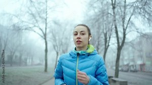 slow motion young sporty woman running with Wireless Earphones. slow motion young sporty woman in blue down jacket running along the alley at foggy winter day and uses Wireless Tws Earphones