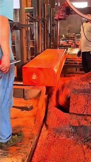 cutting a large reddish wooden log using a giant industrial bandsaw machine in a sawmill factory