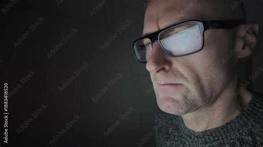 Man sat in dark room using a computer, refection in glasses. Hacker.