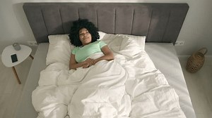 Beautiful sleeping woman of color. Top down morning in bedroom, Health and rest concept. Young African American woman lying in bed and opening her eyes looking at bright sun light lighted up bedroom