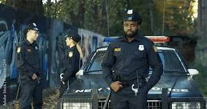 Portrait of serious African American handsome policeman in uniform looking at camera outdoor. Police officer standing and leaning on car. Caucasian colleagues talking on background.