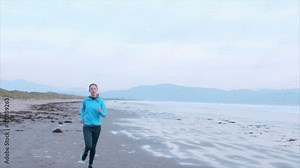 Lockdown video of determined woman jogging on beach. Beautiful female is in sportswear. Tired woman taking break after running. Scenic view of mountains and sea is in background.