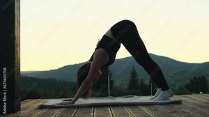 Young athletic woman doing yoga on wooden terrace with mountains view. Female silhouette meditating and practicing yoga poses at dusk. Nature landscape