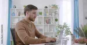 Lateral dolly of handsome and cheerful caucasian brunette man typing on a laptop. Young male professional writing an important email from his home. Remote working day light.