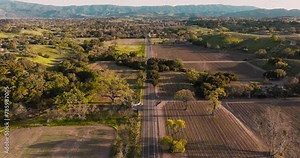 Drone Over Farm Fields in Santa Ynez California, Cars Driving Down Wine Country Road at Golden Hour with Mountains on Horizon