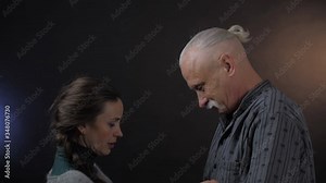 indignant pretty woman with dark hair touches pensioner head shirt and resents against dark background closeup. People emotion