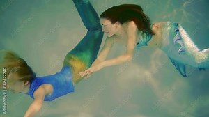 two actresses is wearing fake mermaids costumes and floating under water in a pool, posing and playing