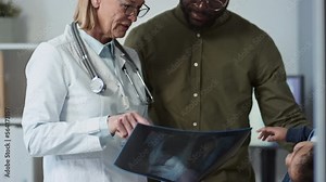 Tilt down and up of female radiologist in lab coat commenting lung x-ray result during conversation with young African American man visiting clinics with his little son Stock Video