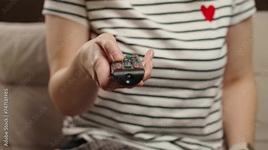 Woman holds TV remote control. Female is sitting on sofa in living room and switching channels on the TV. Concept of entertainment and lazy weekends