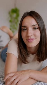 Beautiful young woman smiling lying on bed at home. Joyful portrait of pretty millennial female with toothy smile looking at camera. Vertical screen HD video footage.