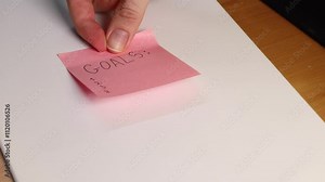 Hand sticking pink sticky note with word “GOALS” written on it. Symbol of planning and motivation. Close-up highlights goal-setting concept and future aspirations.