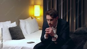 upset businessman sitting on bed with clenched hands in hotel room