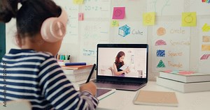 Asian toddler girl with sweater wear headphone sit front of desk with notepad use magic pen focus on writing do homework listen teacher online learning english course from laptop screen at home.