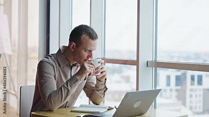 Calm relaxed man sit in front of his computer with a cup in hands. Thoughtful man looks into window and then straight ahead.