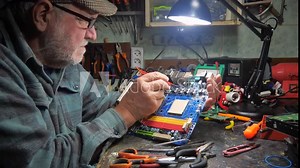 An old master repairs computer components in a workshop. Using a soldering iron and other tools, he repairs and adjusts broken electronics.