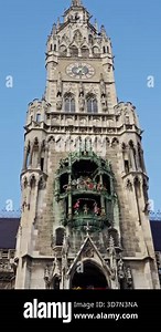 architectural facade under bright blue sky, sunlit stonework and floral displays adorn historic clock building, vibrant flags and sculpted statues accentuate grand gothic clock tower scene. Munich. 4k