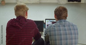 Rear view of two outsource software developer looking at computer screen and discussing source codes while working on project at creative office. IT programmers with laptops typing lines of HTML code.