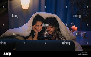 Multiracial couple man and woman shocked watching horror film movie scared afraid frightened watch video with mobile phone hiding under blanket cover Stock Photo - Alamy