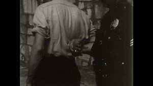 1940s: Police officer uses leather strap and baton to restrain the hands of a man. Officer twists man's wrist and guides him along sidewalk with baton.