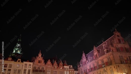 Low-angle night view of the historic Grote Markt buildings, highlighting their illuminated facades and a prominent clock tower against a vast dark sky. Creates sense of grandeur.