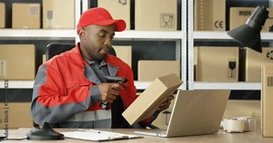 African American mailman in uniform working at laptop computer in post office store with parcels. Postman scanning carton box with scanner, registering it and filling in invoice document.
