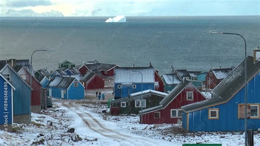 Colorful houses sit on a rocky shore with snow and ice in Greenland. The ocean waves crash against the land as clouds drift above. It shows the simple life in this remote location.