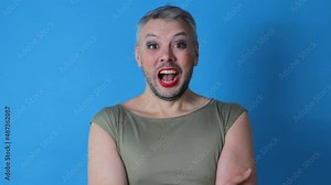 a gay man with makeup on his face opens his mouth wide in surprise against an isolated blue background. LGBT. Transgender