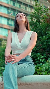 VERTICAL VIDEO: Young woman with freckles and dark loose hair wearing white top enjoying the view of modern city. Beautiful girl with long eyelashes looks at the upper floors of building