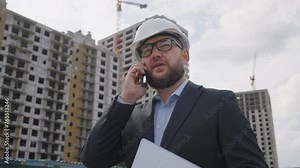 Construction project manager in suit and hardhat walking and talking on phone, with laptop in hand, at bustling construction site