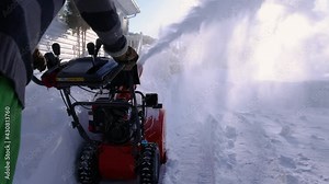 View from behind of a bright orange mechanical snowplough being pushed by a young man to clear a path from the abundant fresh snow recently fallen.