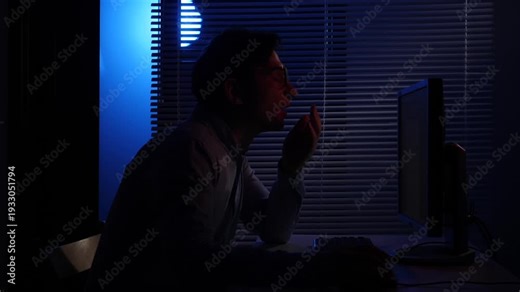 Overworked man yawning while staying late at night in a dark office, managing a deadline and facing fatigue from long hours of computer work and overtime