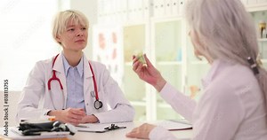 Blonde doctor in work uniform gives bottle of pills to sick elderly woman sitting at table in light office. Specialist prescribes medication slow motion